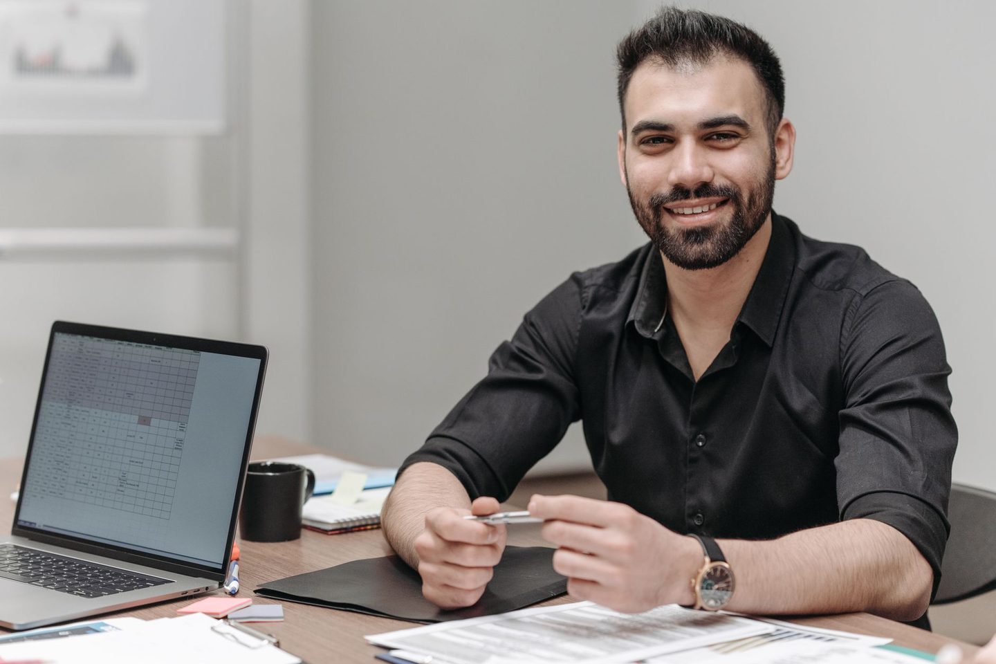 A smiling finance manager at a desk with a spreadsheet open on a laptop and a schedule of papers in front of him